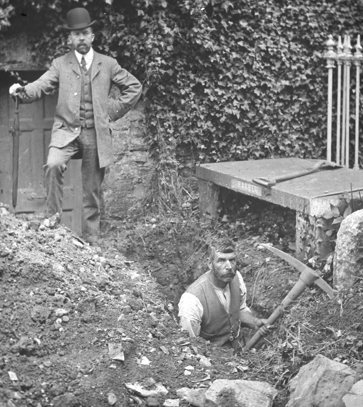 A man stands over the excavated grave while a labourer stops his work for the photograph.