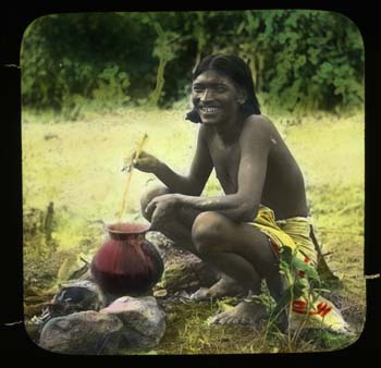 Man, cooking pot on open fire, beside thatched dwelling.  This man features in some of the other lantern slides